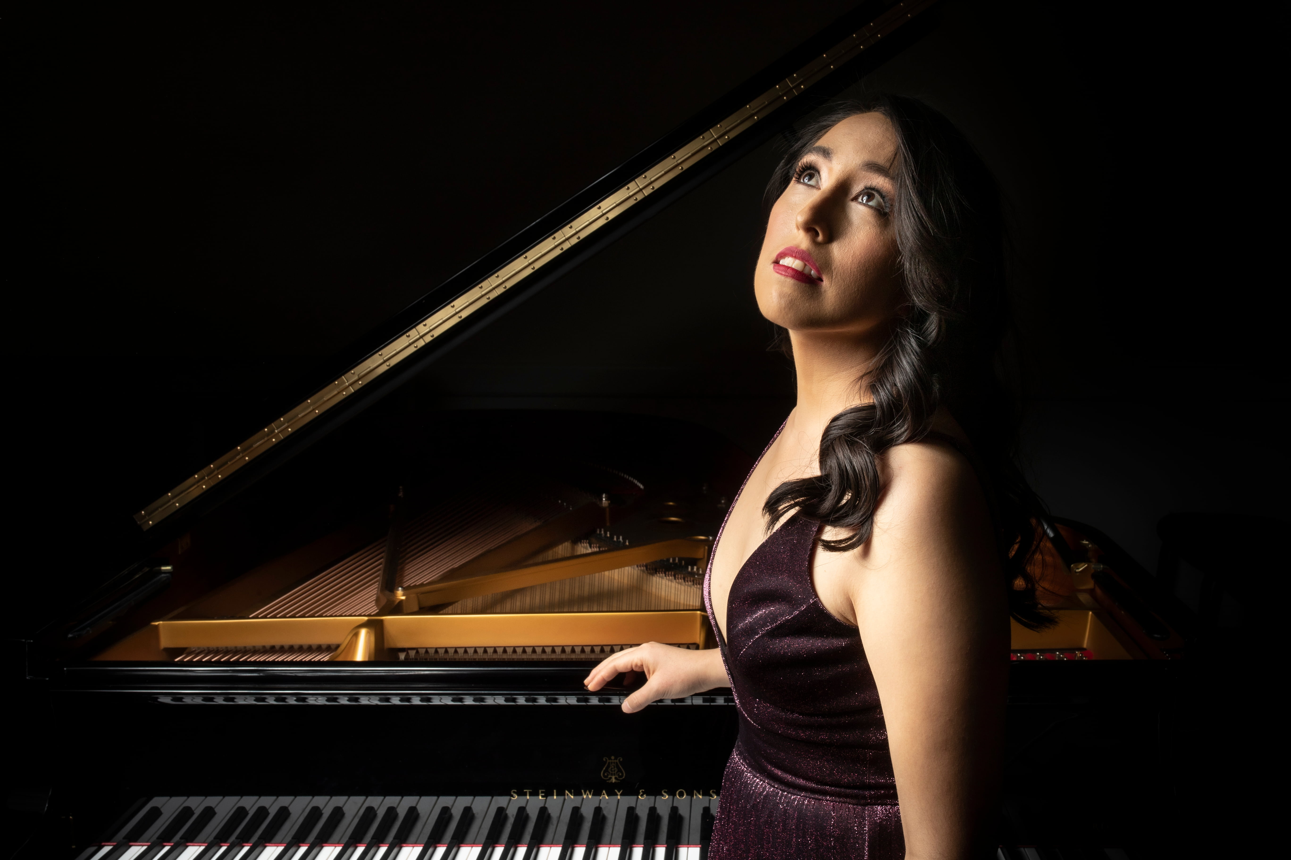 Woman with long dark hair standing in front of grand piano in formal wear looking dramatically up and off camera