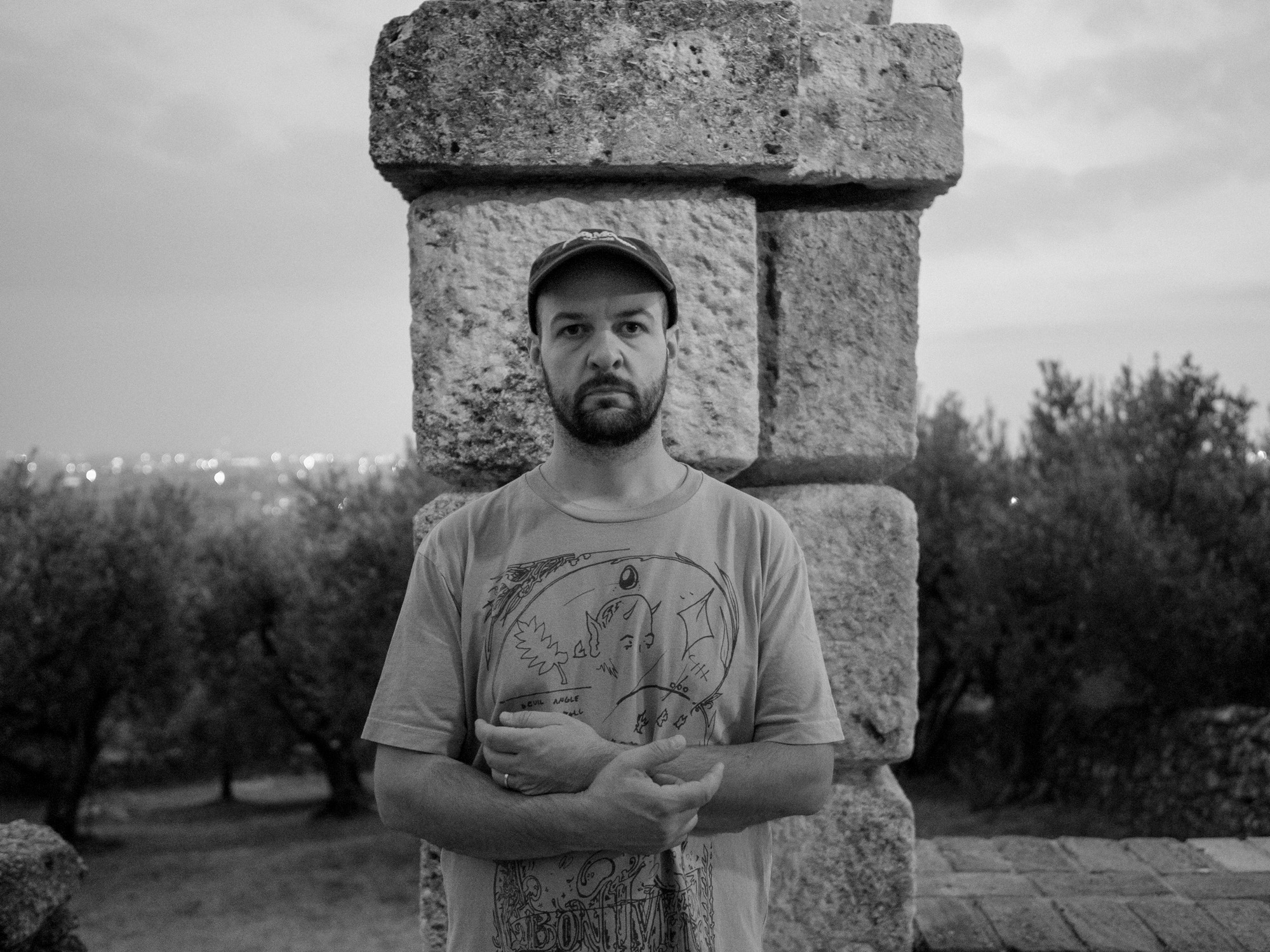 Man wearing t-shirt and hat standing in front of a stone pillar with arms folded in front of him