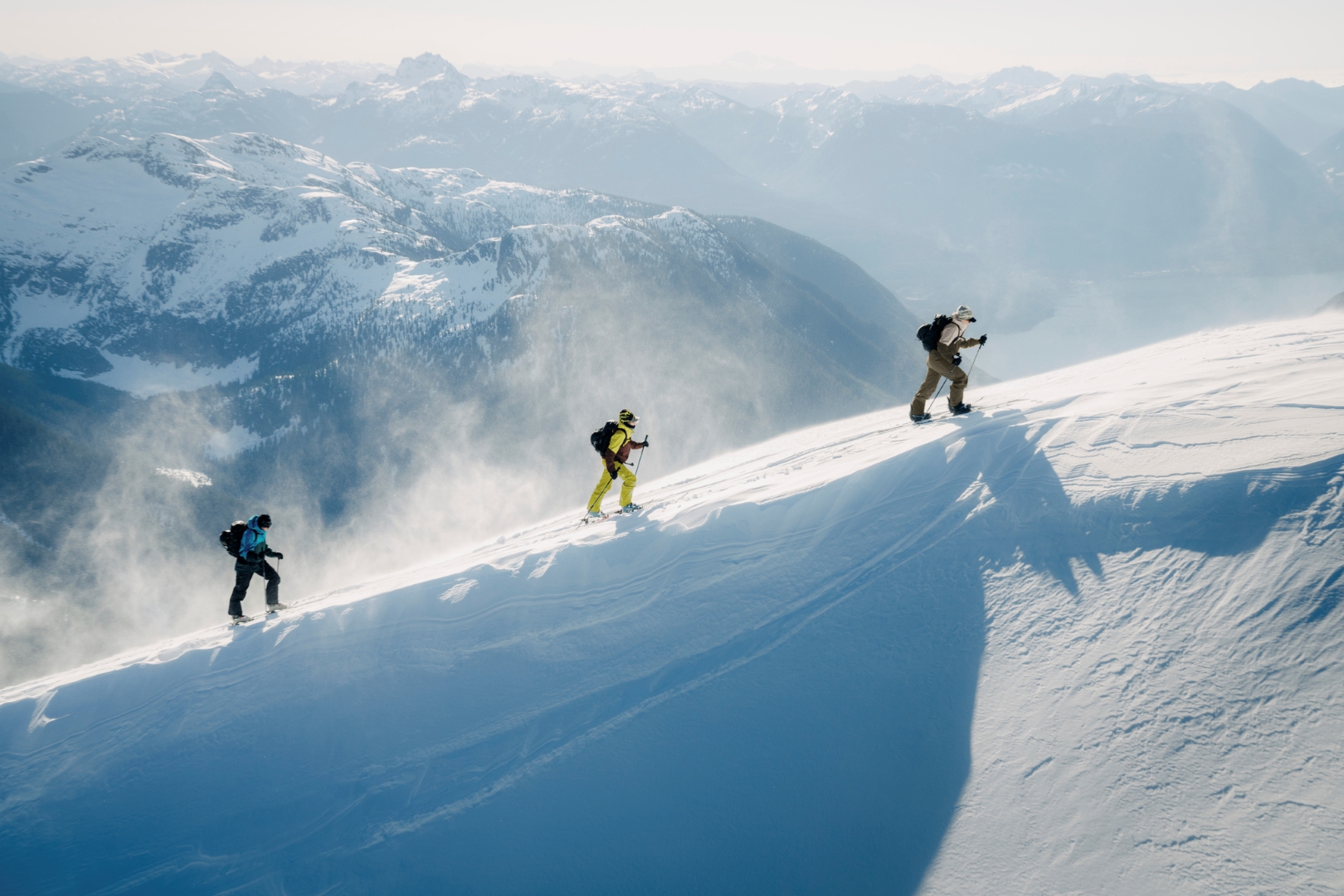 Two skiers and a snowboarder tour up a snowy alpine ridge in full shell jackets and pants. They are in the Tantalus range of the British Columbia Coast Mountains. 