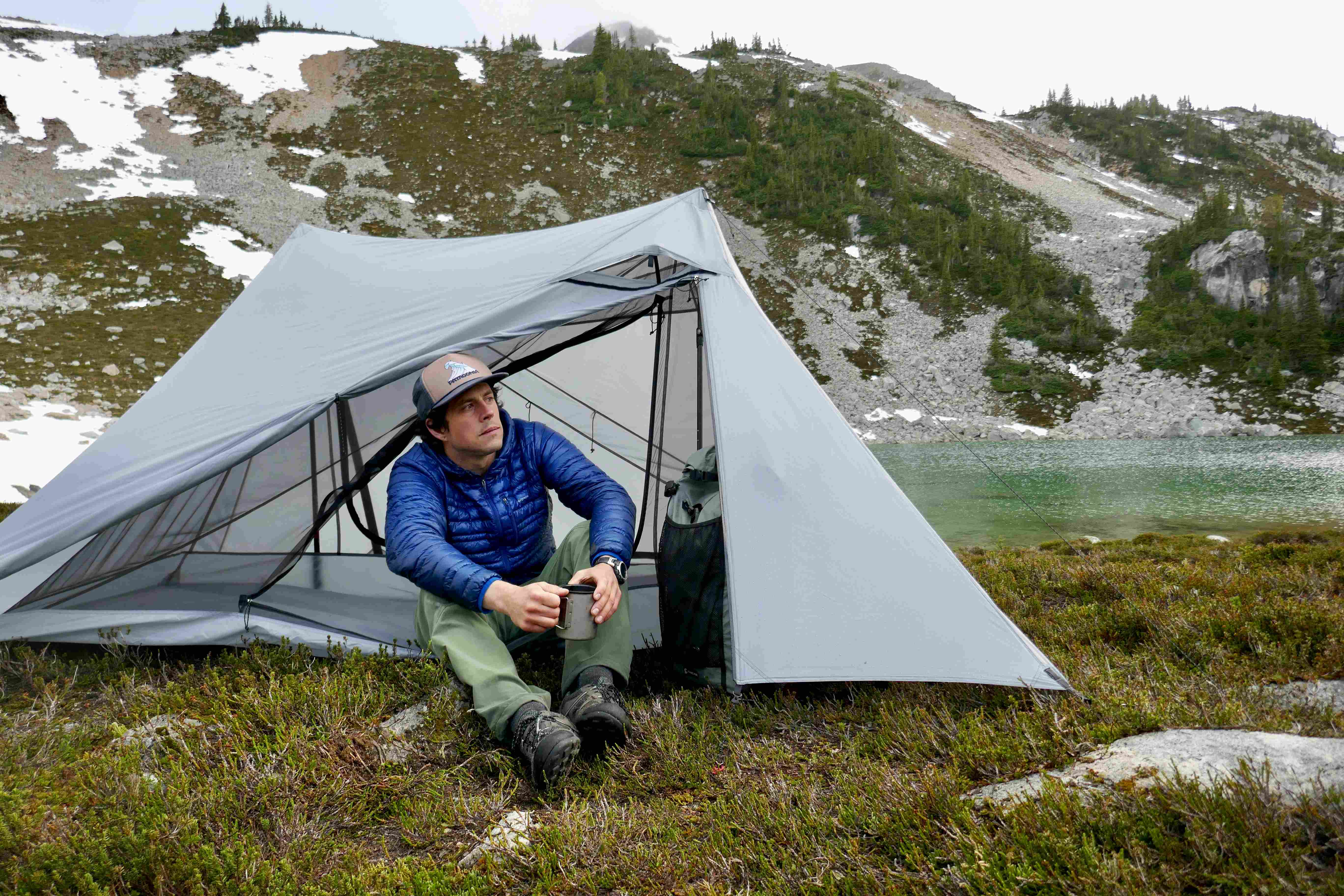A man holding a coffee mug sits in a unique looking dyneema tent in the mountains. The tent is on a patch of grass by a 