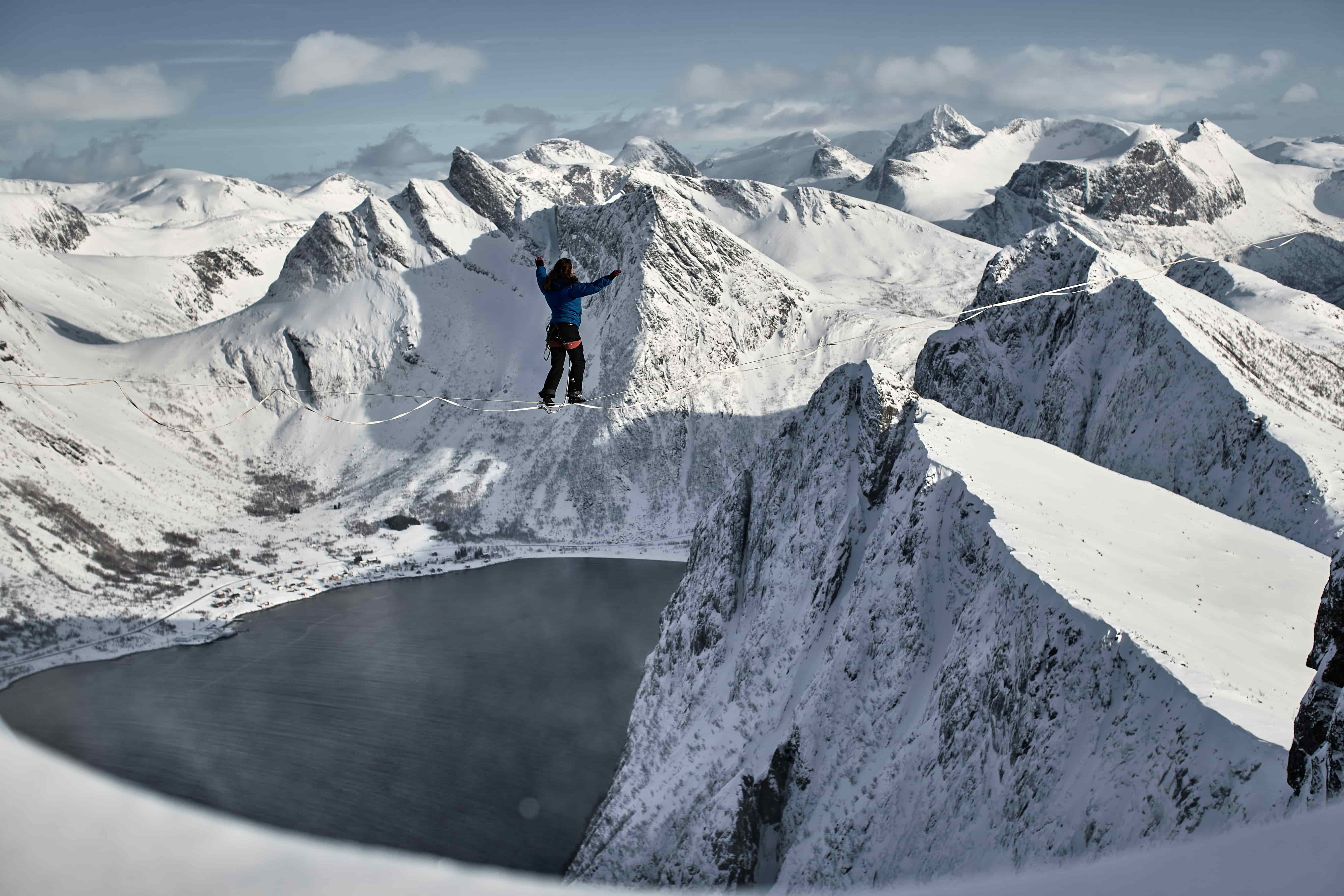 An athlete is walking on a slackline amongst snowcapped mountain peaks.