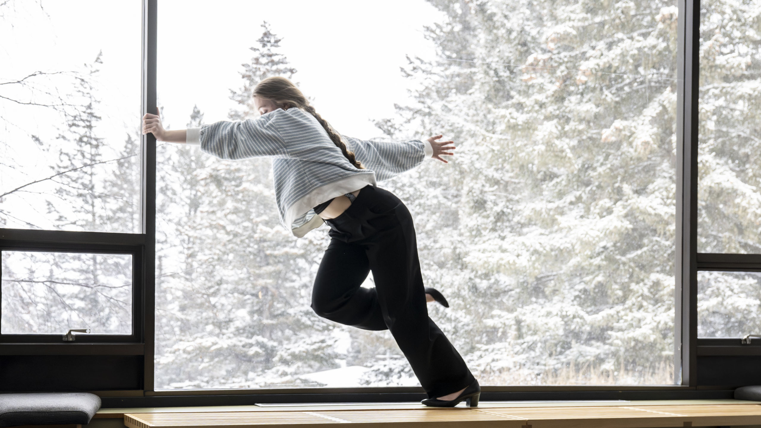 A dancer is stretching in front of a window with snowy trees in the background