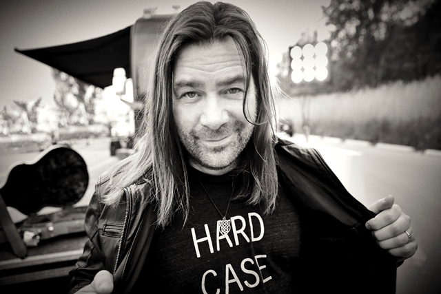 Alan Doyle holds the left and right sippers of his black leather jacket open displaying a t-shirt with the words "HARD C