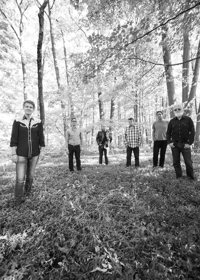 The six members of Blue Rodeo stand in the grass beneath a maple forest.