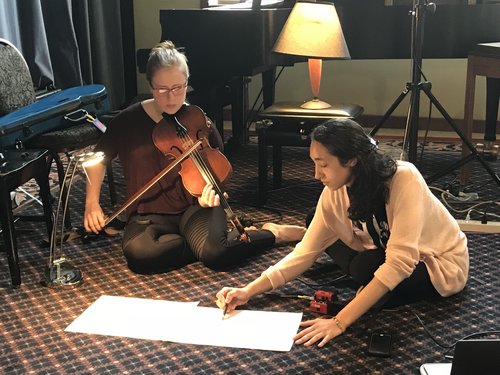 Two participants are seated on the floor. One annotates music while the other looks on holding a violin at the ready. 