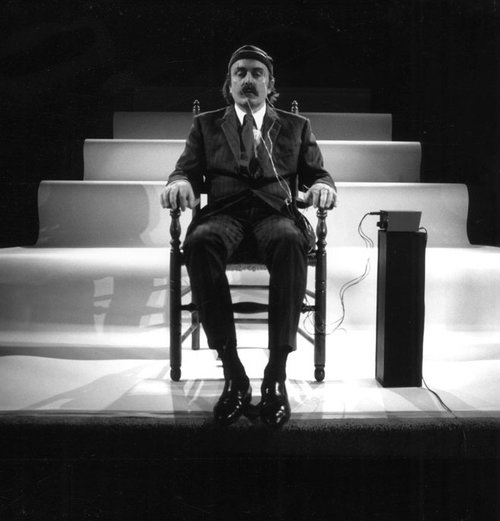 A black and white photo of musician Alvin Lucier sitting in a wood chair with what appear to be electric pulse nodes att