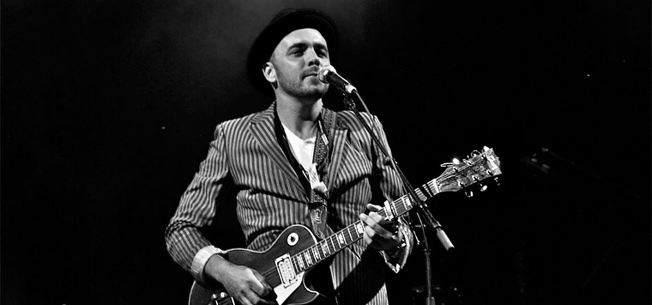 Hawksley Workman plays guitar before a mic in a black and white photo.