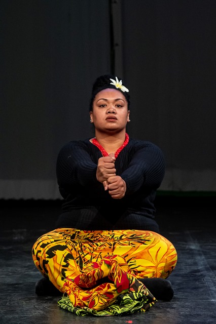 An indigenous woman sits on the ground with her hands in stacked fists held in front of her. 