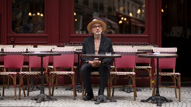 A man with glasses and a hat sits in a black suit with no shirt at a cafe patio setting. 