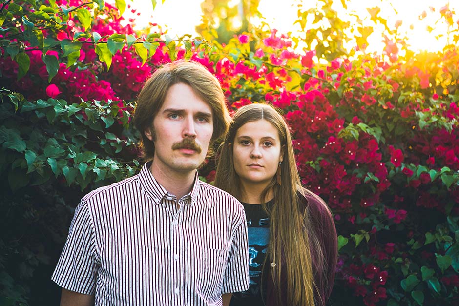 Kacy and Clayton are standing outdoors in front of a colorful hedge of green leaves and fuschia flowers. 