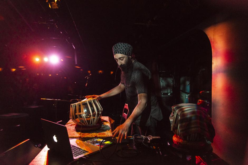 A bearded man stands over a unique drum with electronics beside him.