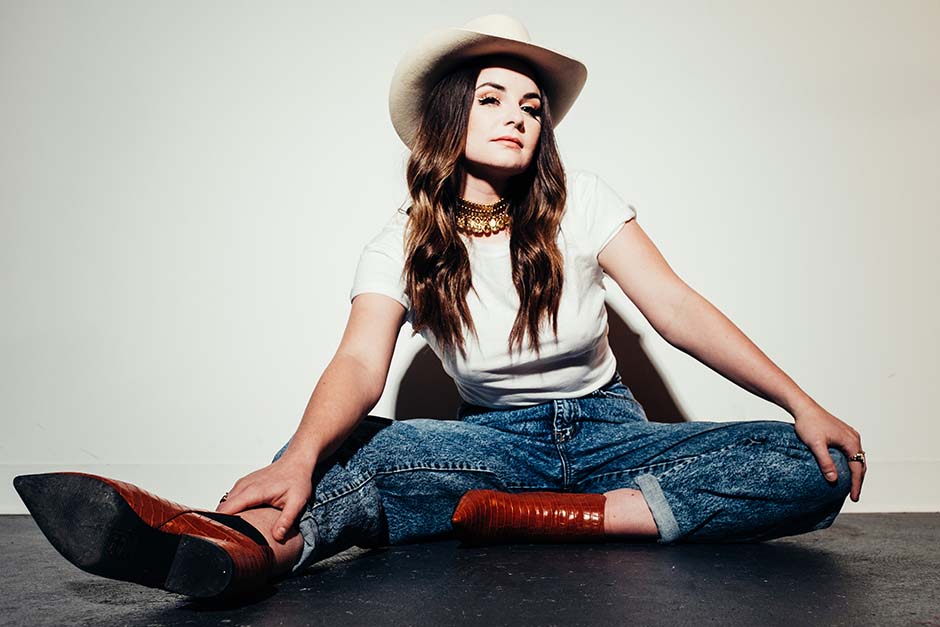 Mariya Stokes is wearing brown boots, blue jeans, white t-shirt, and a white cowboy hat. She is seated on the ground in 