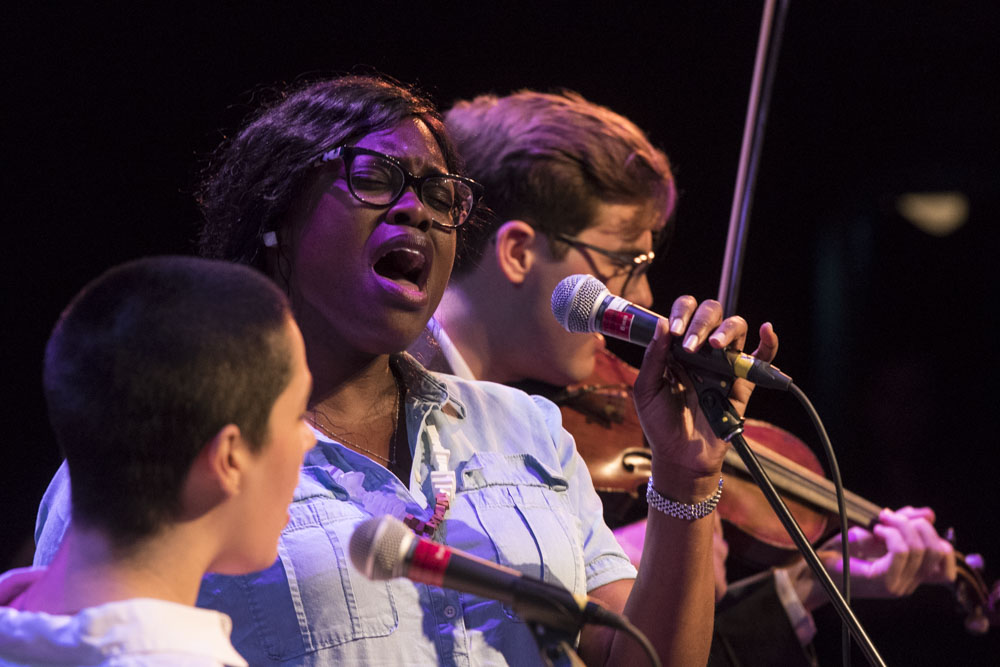 Two Banff Centre music participants sing into a microphone with a violinist behind them.