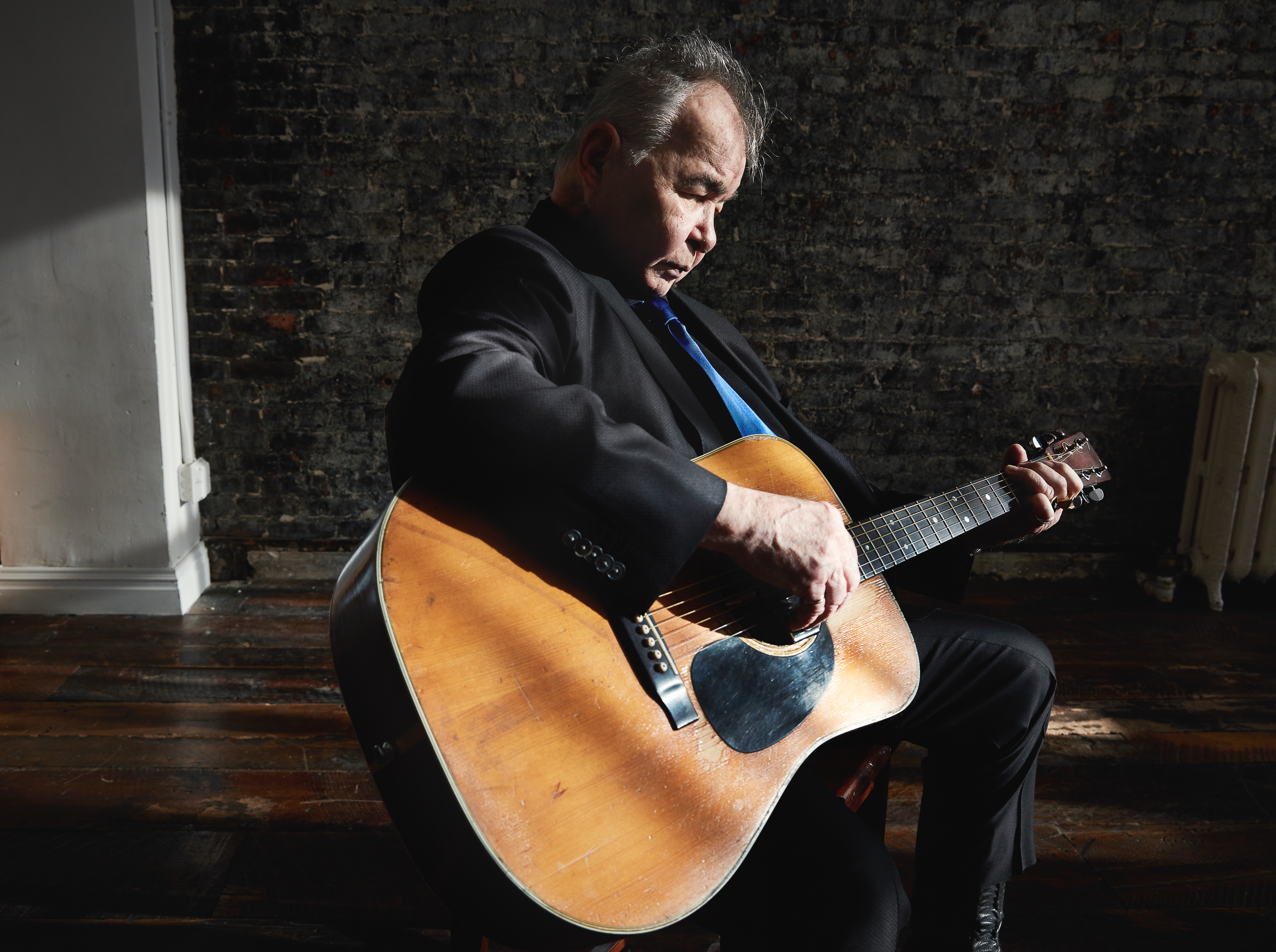 John Prine sits playing an acoustic guitar in an informal space.