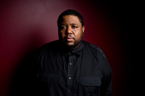 Tyshawn Sorey stands before a red wall wearing a black shirt and has his arms behind his back.