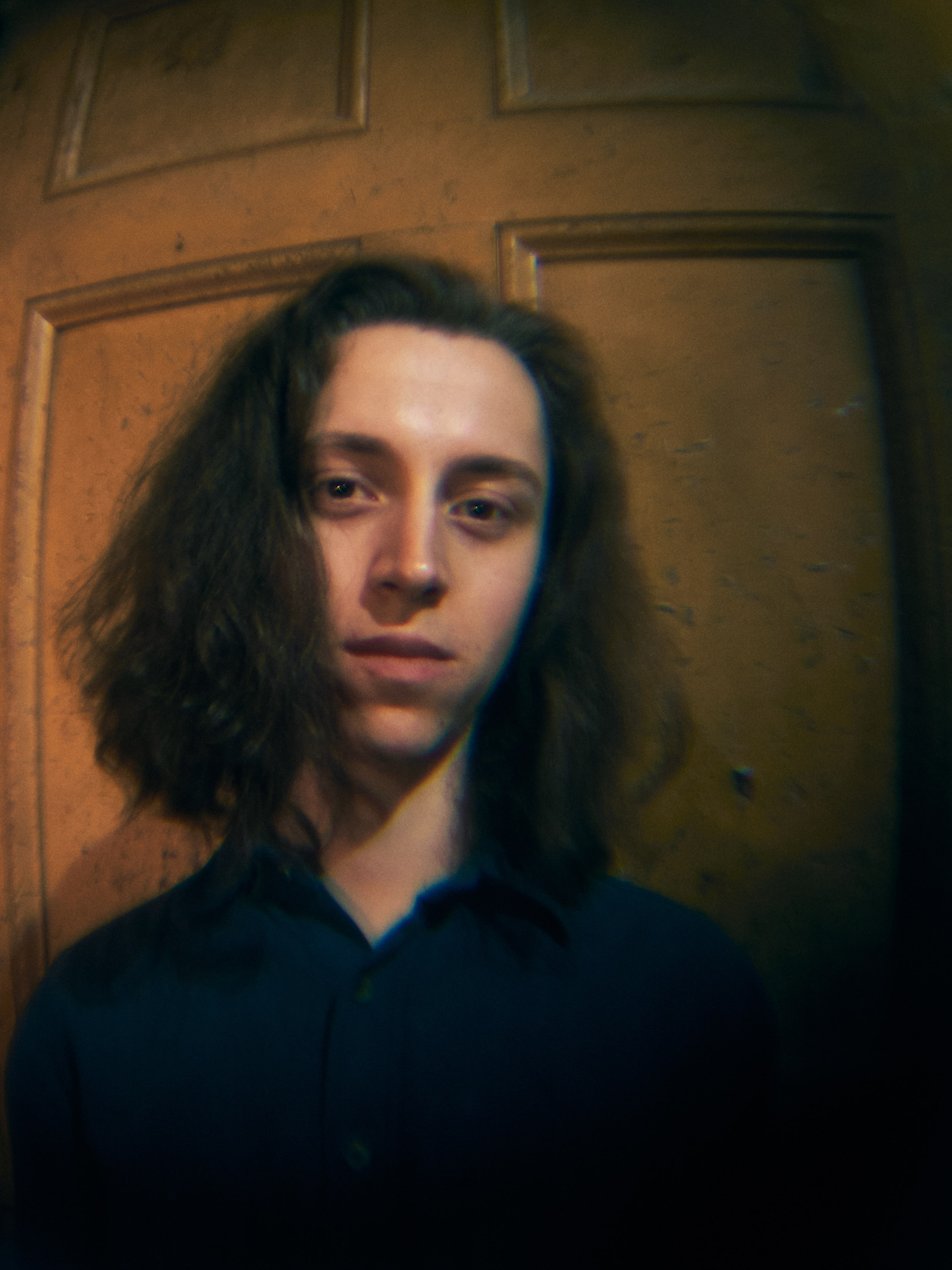 Man with long shoulder length hair standing against door looking at camera