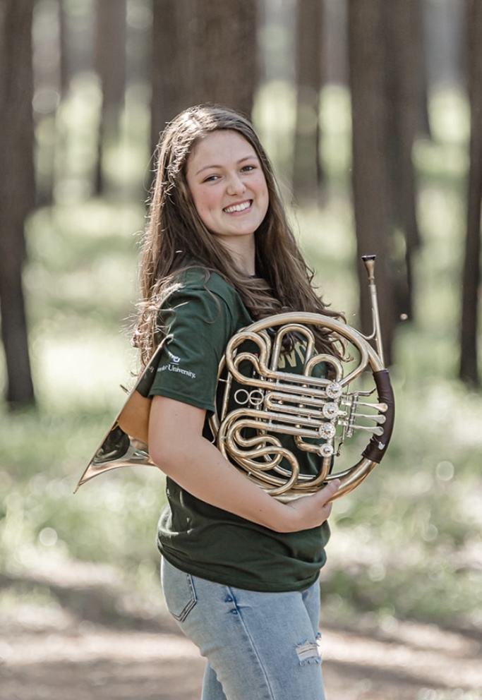 Woman standing in forest holding french horn and smiling at camera