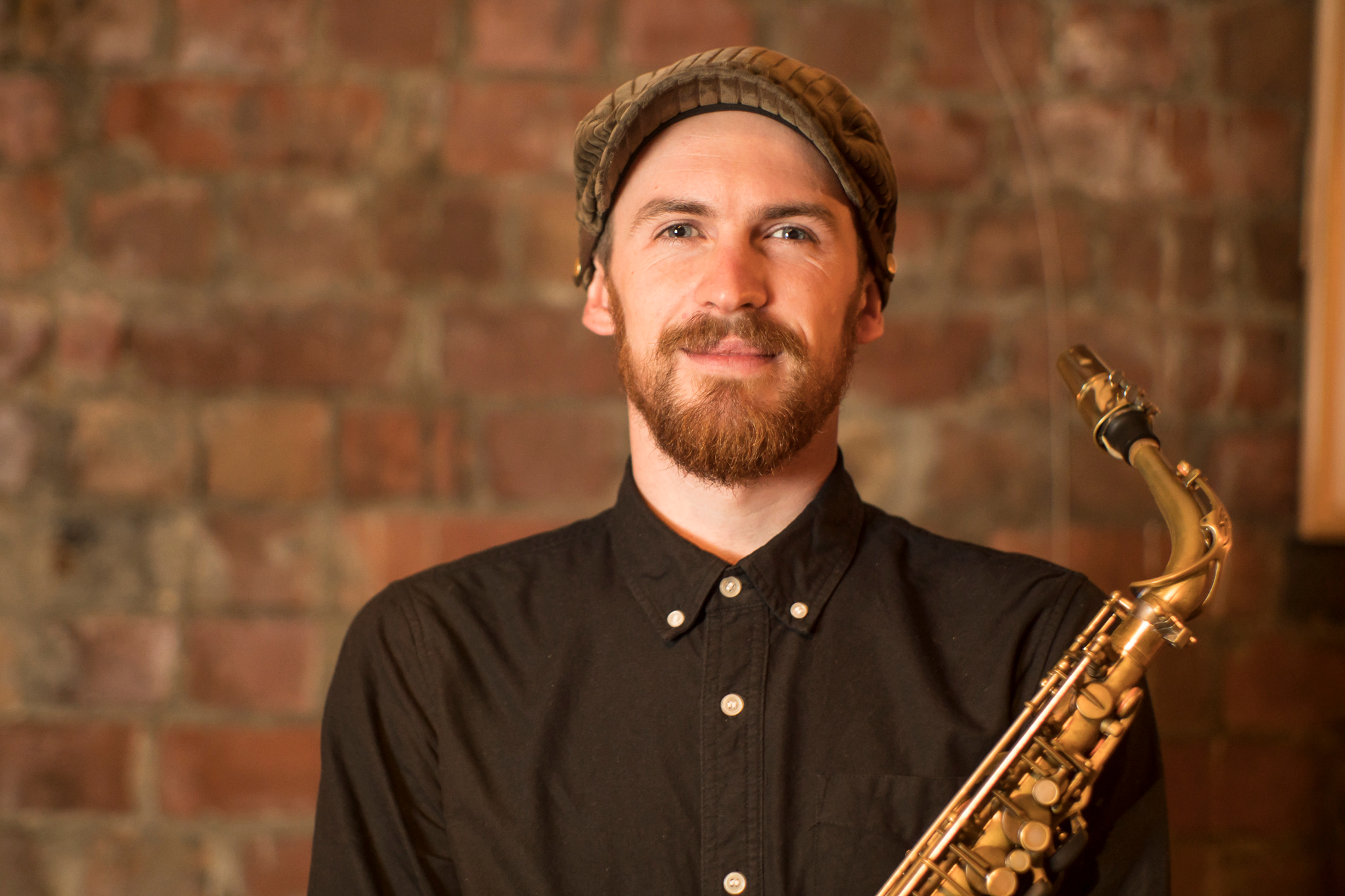 Man wearing cap and black button up smiling at camera and holding a saxophone in front of him