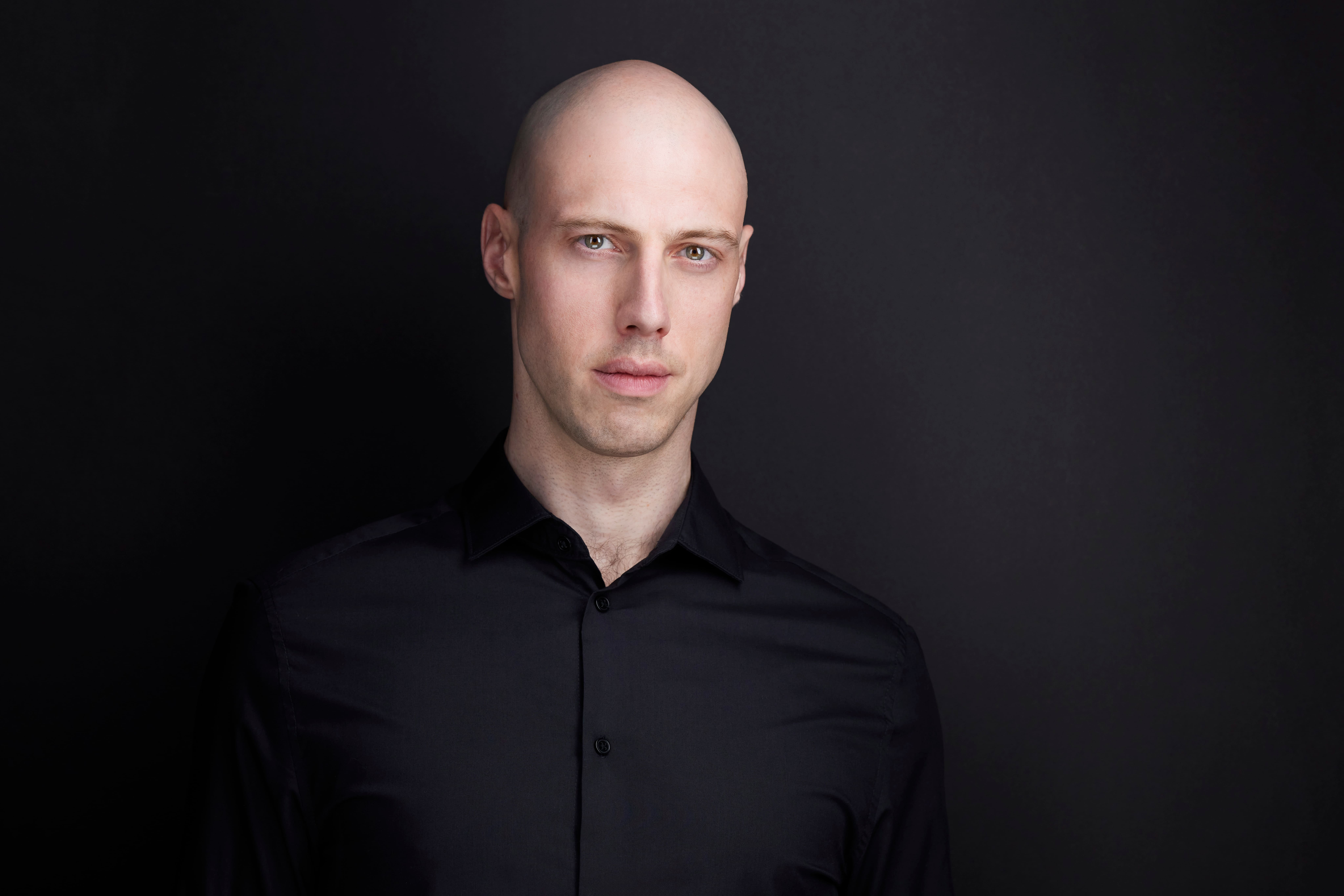 Man in black shirt standing in front of black wall staring intensely at camera