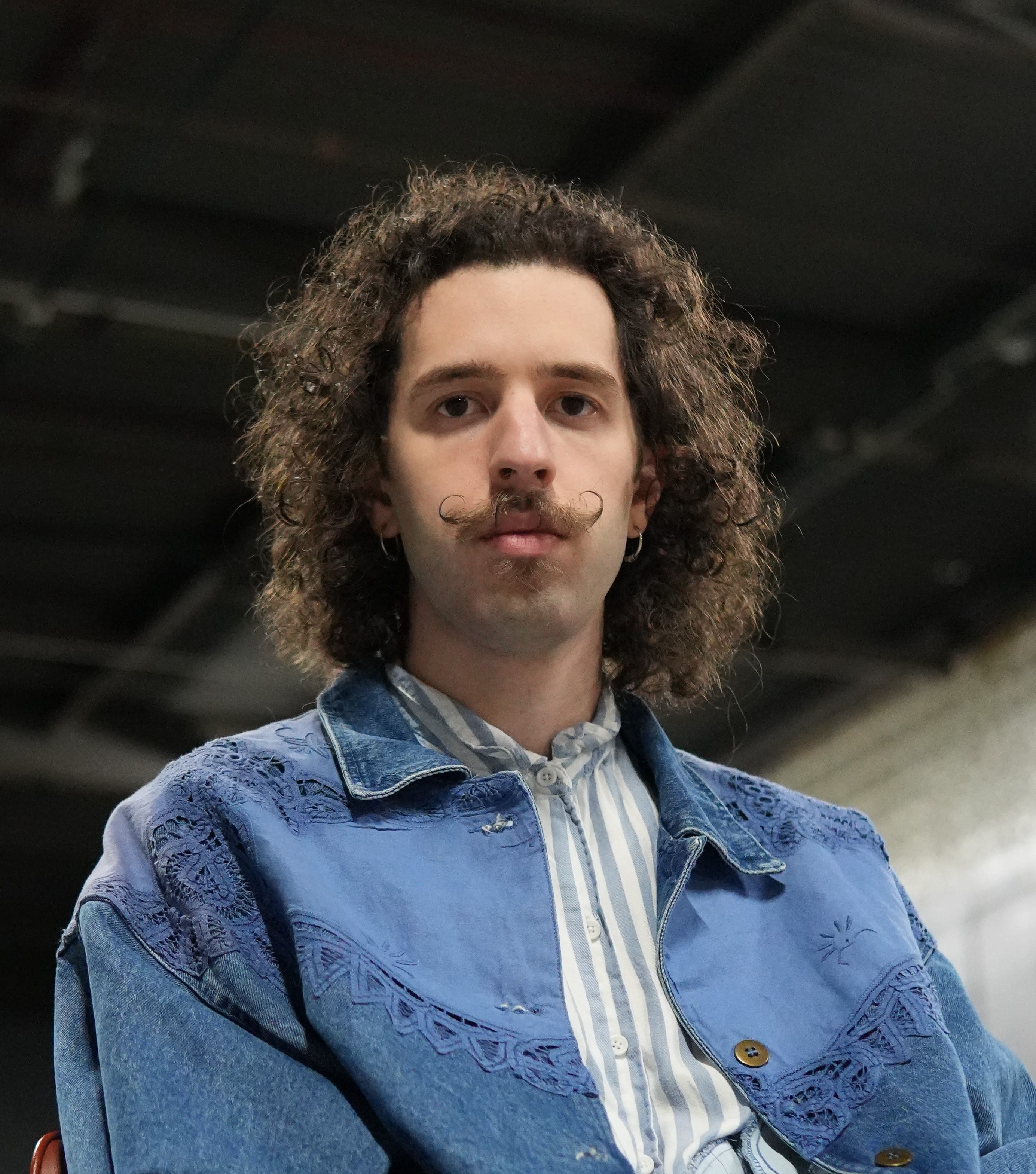 Man with shoulder length curly hair looking at camera wearing a jean jacket and a distinct moustache