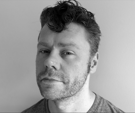 Man with short curly hair smiling slightly at the camera in black and white image