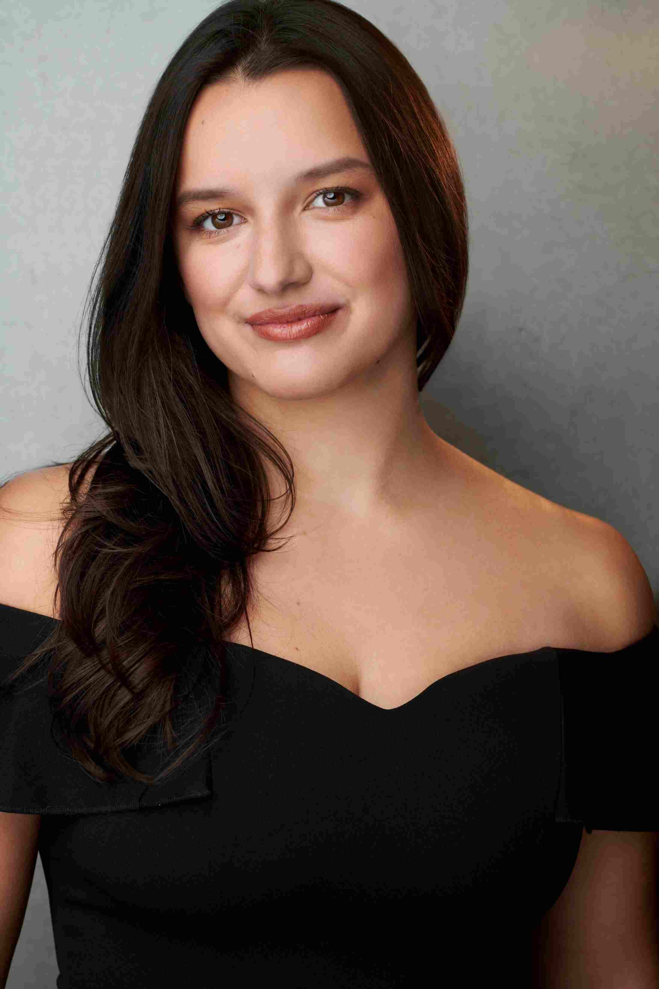 Woman with long black hair flowing over one shoulder smiling at camera in off the shoulder black dress