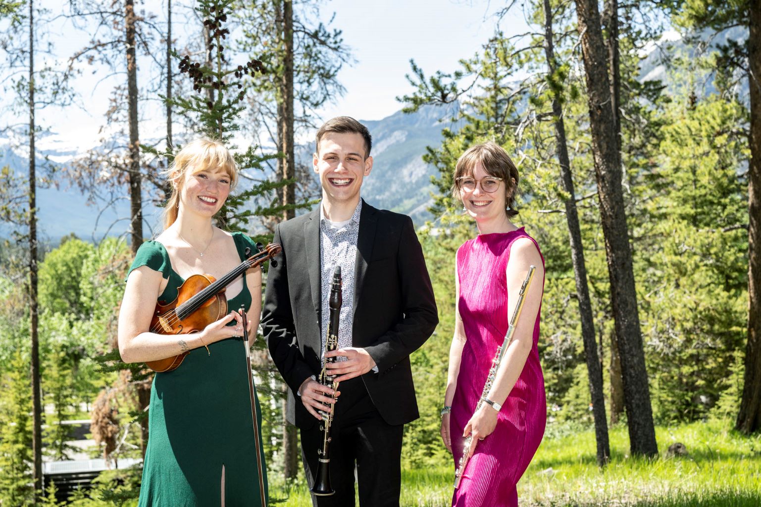 Woman in green dress holding a violin standing beside man in suit with a clarinet in front of him next to a woman in a pink dress holding a flute casually at her side
