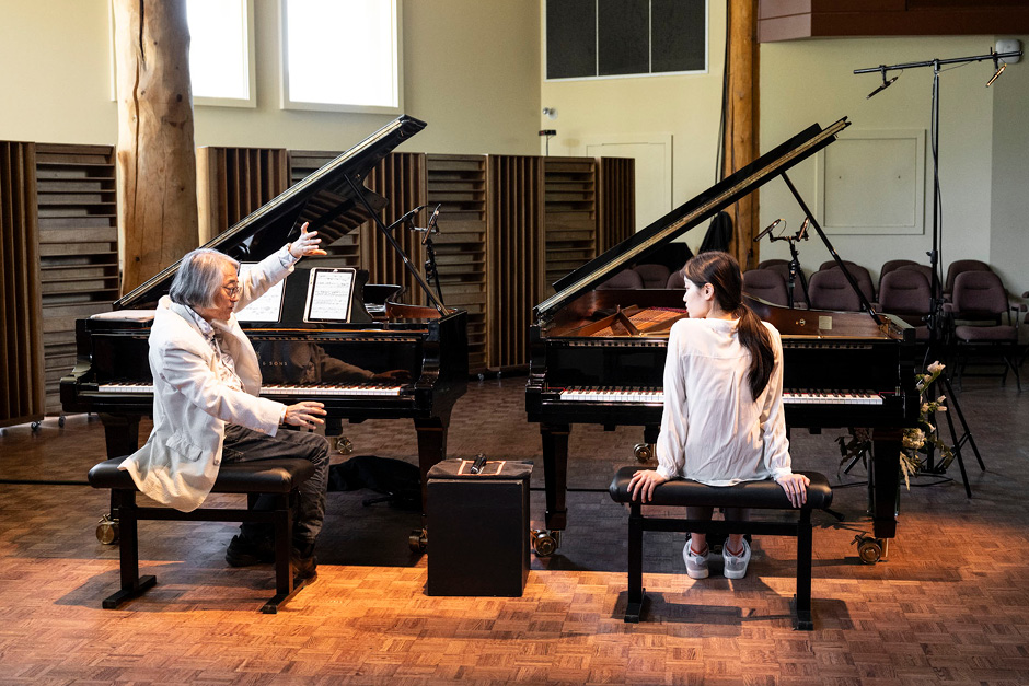 Two people are seated at a pair of grand pianos