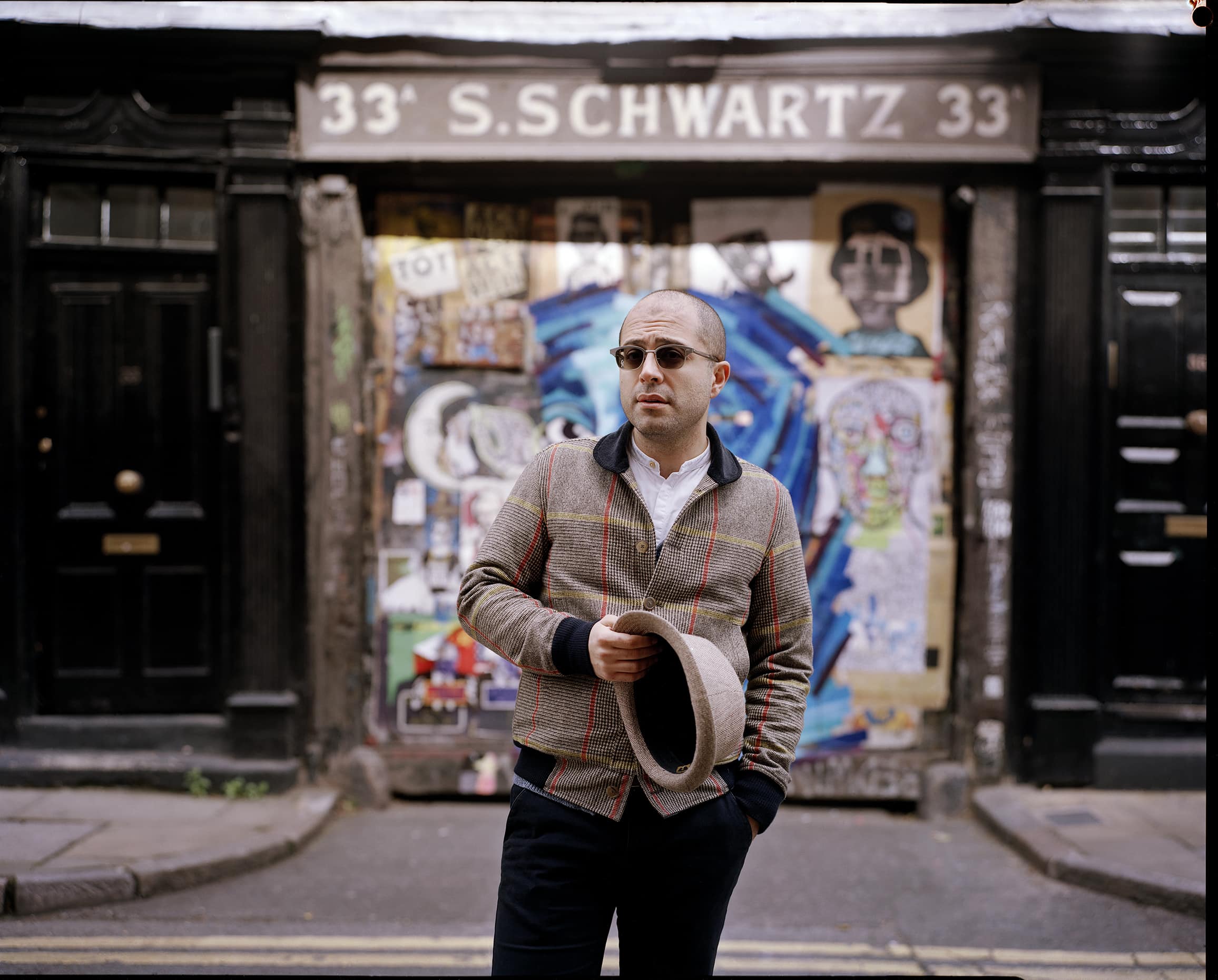 A man wearing sunglasses and holding a hat in hand is standing in front of a graffiti covered wall