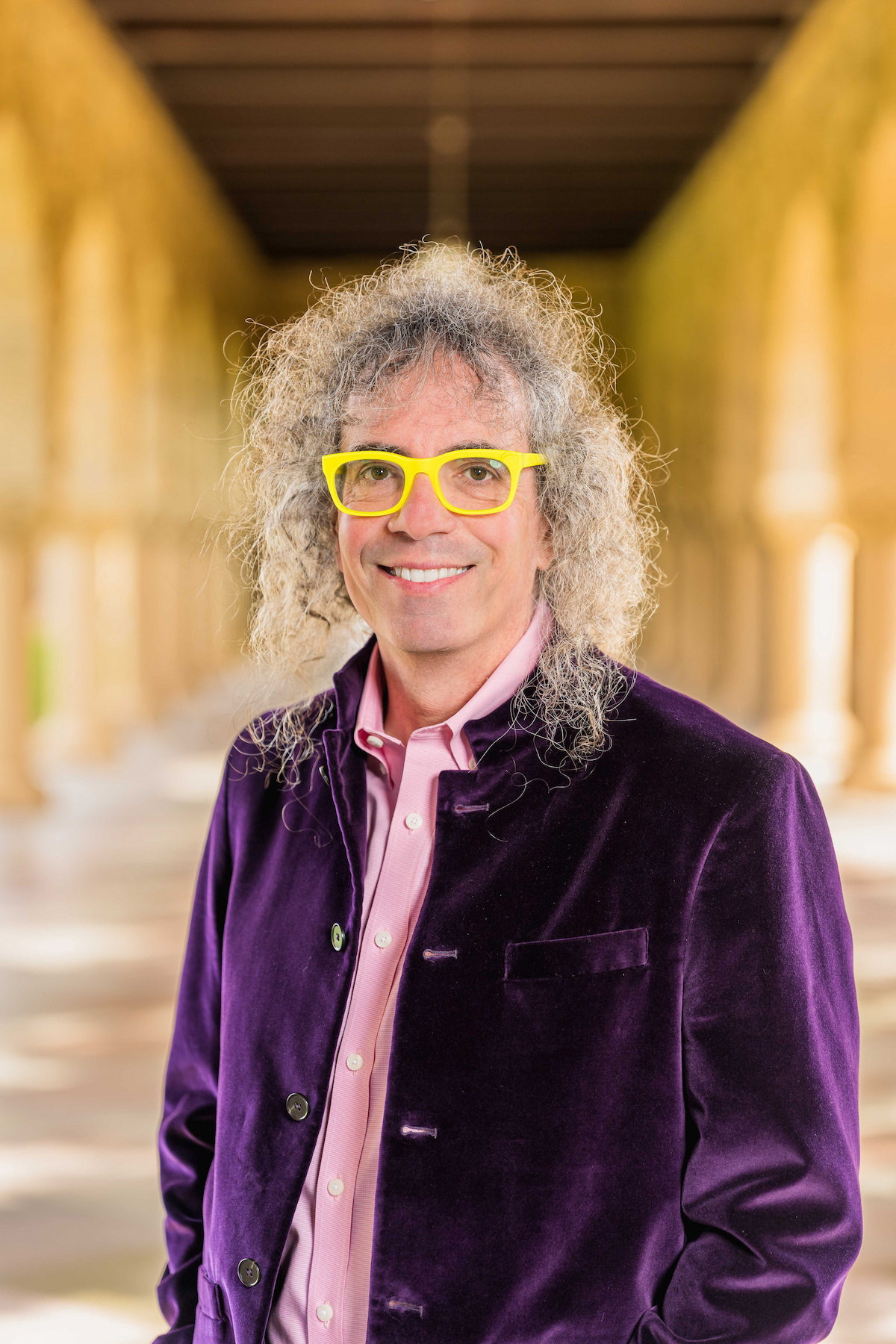 A man wearing a purple jacket and yellow glasses smiles at the camera