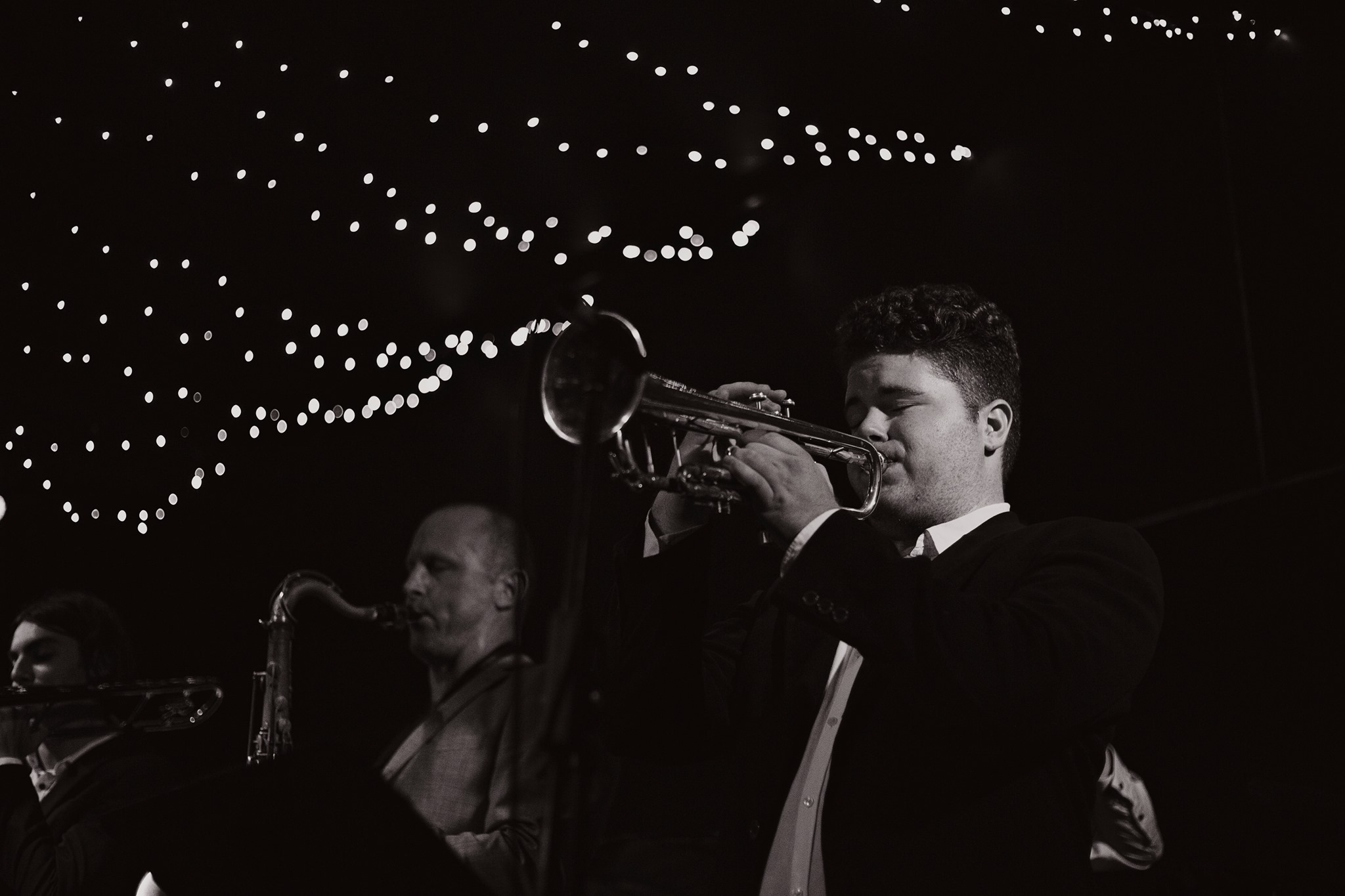 Photo taken at night with man playing trumpet under string of lights