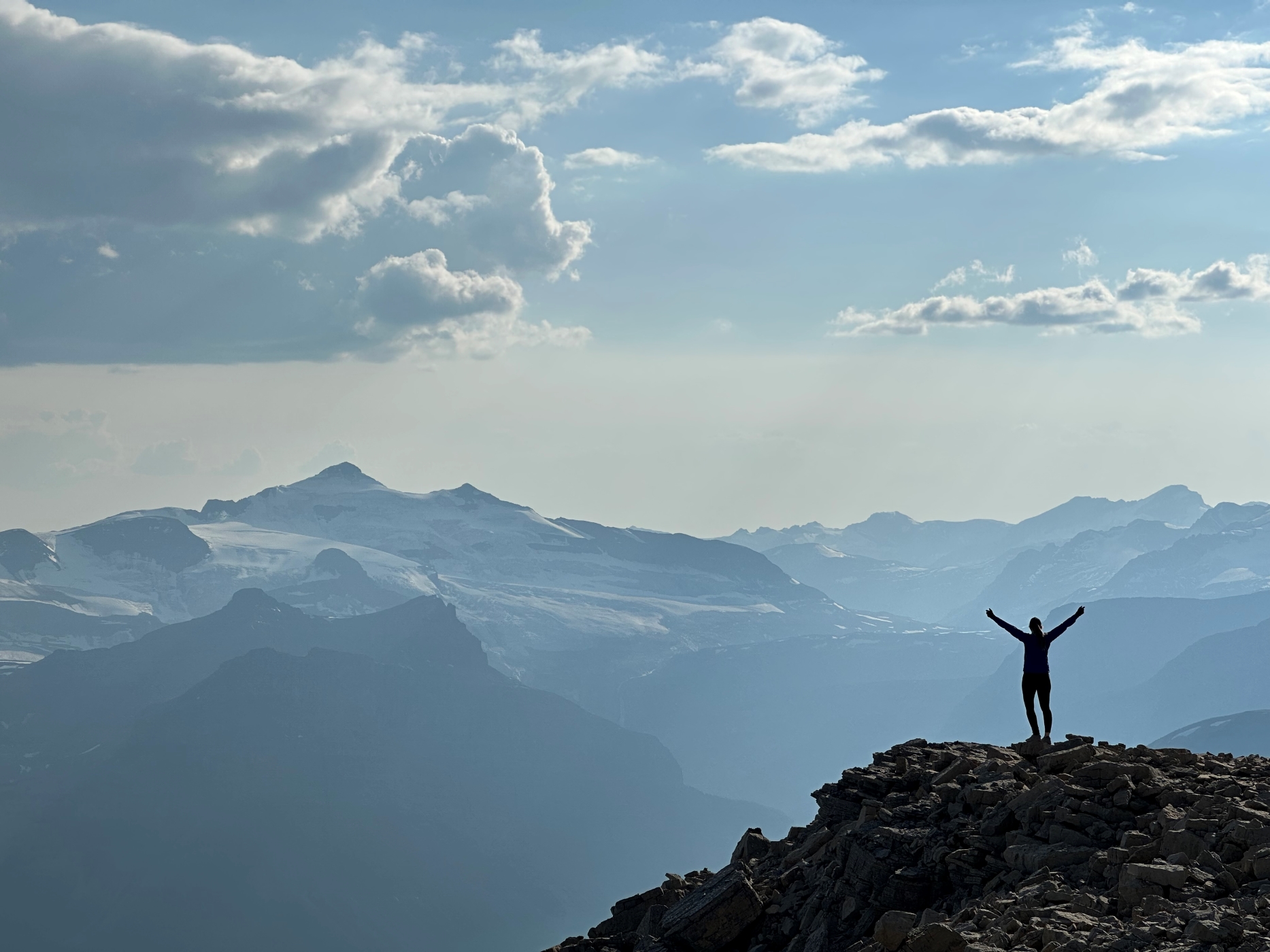 A younf woman stands on a rock outcrop of a peak, her hand extend upwards in a celebratory manner. Glaciers and icefields of the Canadian Rockies can be seen behind her.