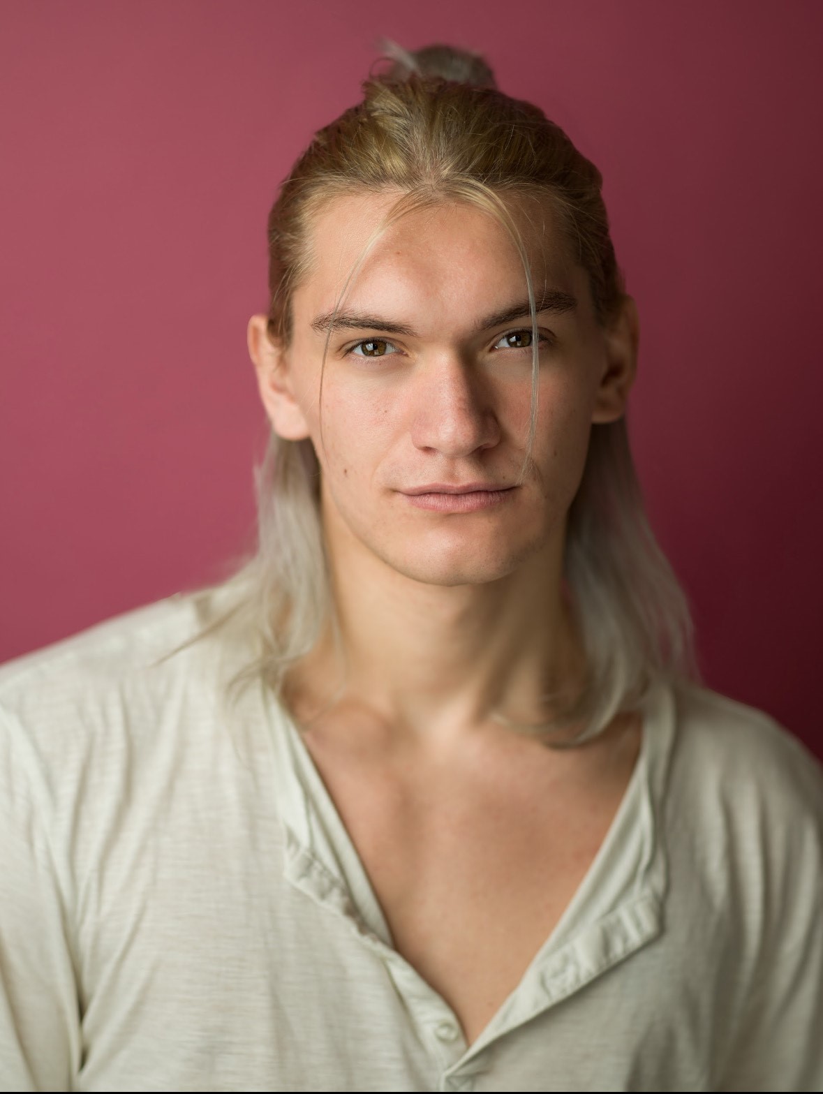 Man with shoulder length blond hair staring at camera with slight smile in front of pink backdrop