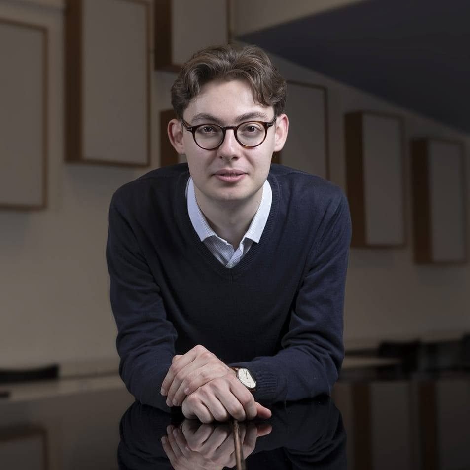 Man with glasses in black sweater and white collared shirt looking at camera