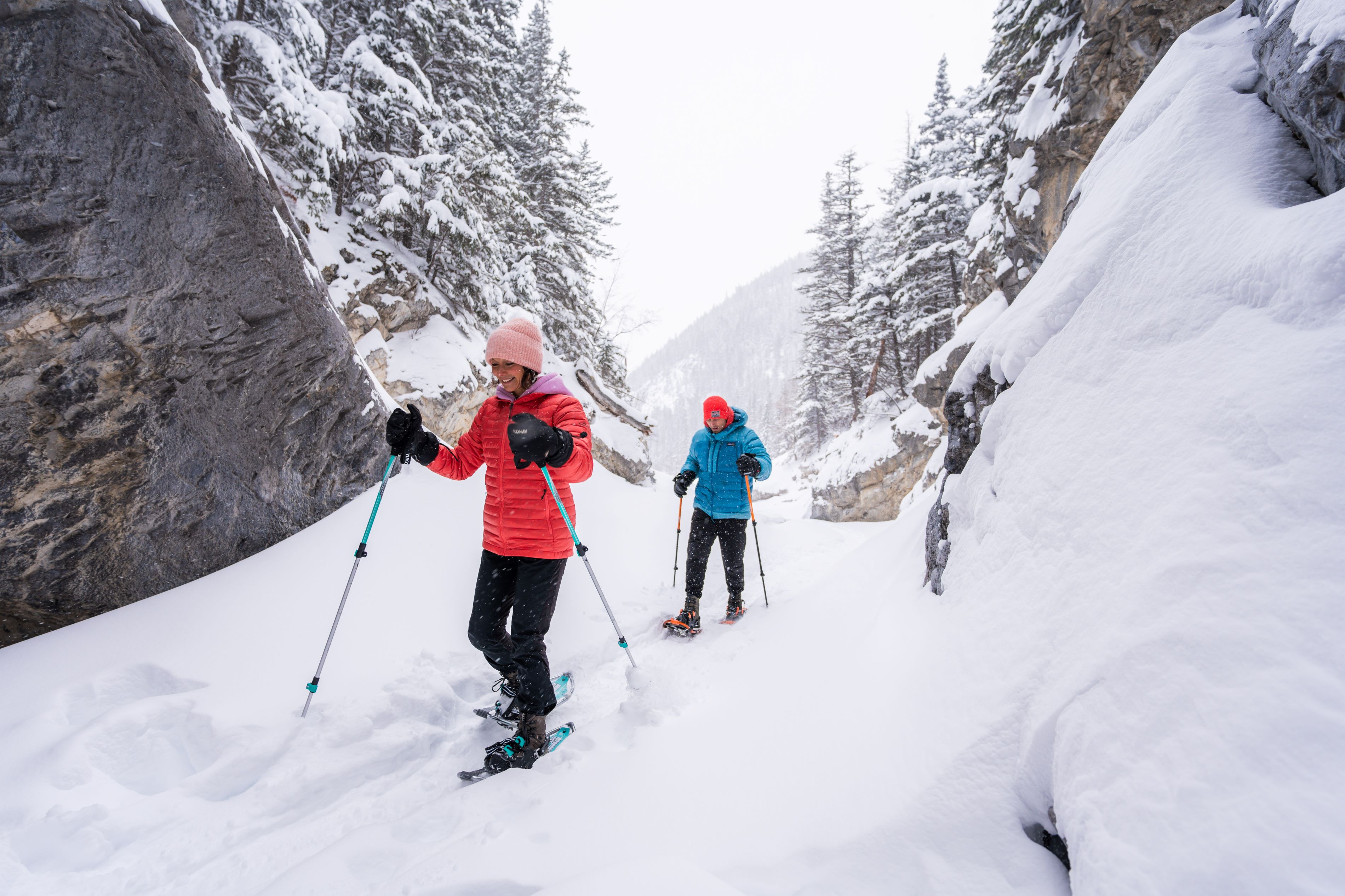 A woman and a man in down jackets, and full winter wear snowshoe up a shallow limestone canyon filled with newly fallen snow.