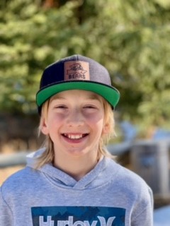 Young blonde boy smiling directly at camera outside wearing a baseball cap
