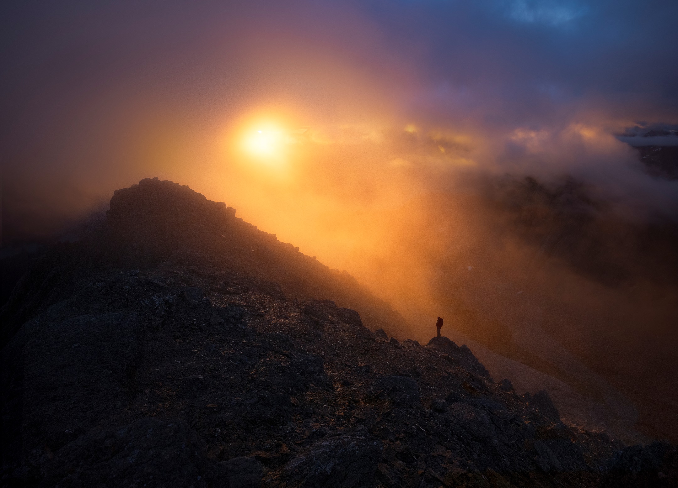 A figure silhouetted by a colourful misty sunrise stands upon a rubbly peak. 