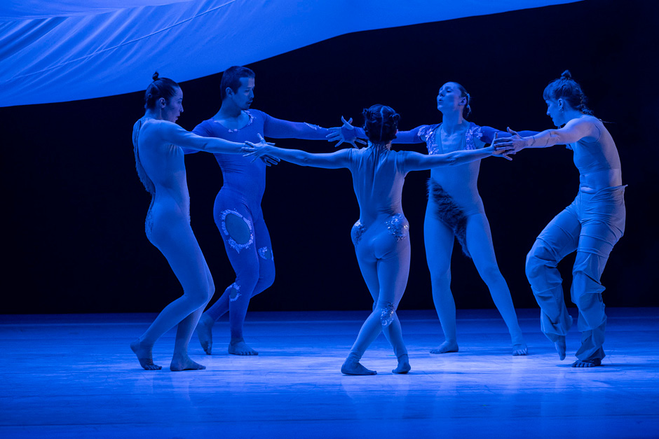 Five dancers are posed in a circle with their arms outstretched and touching, on a stage that is illuminated with blue l