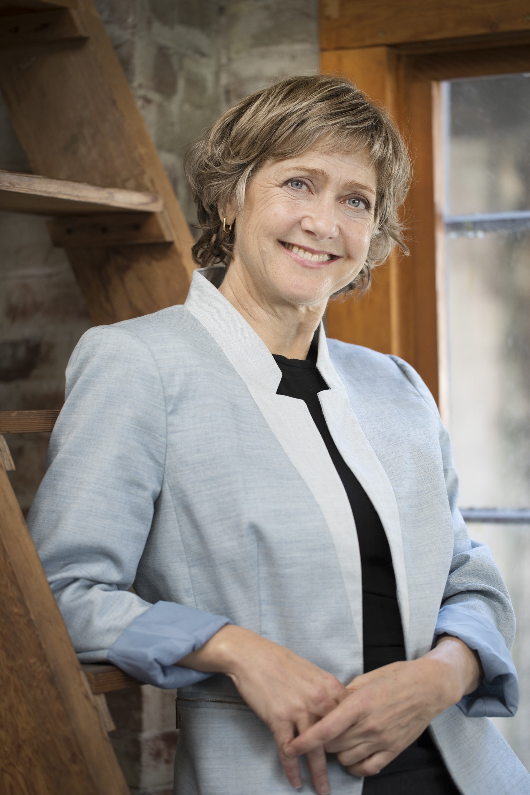 Woman leaning against a wooden ladder smiling broadly at the camera in a blazer with popped collar
