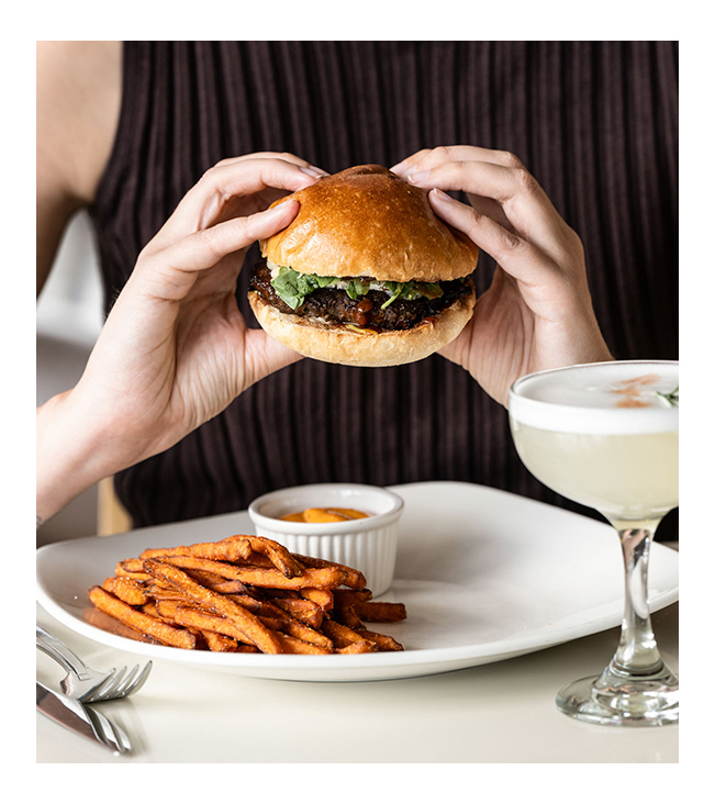 Person holding hamburger with fries and cocktail in the frame.