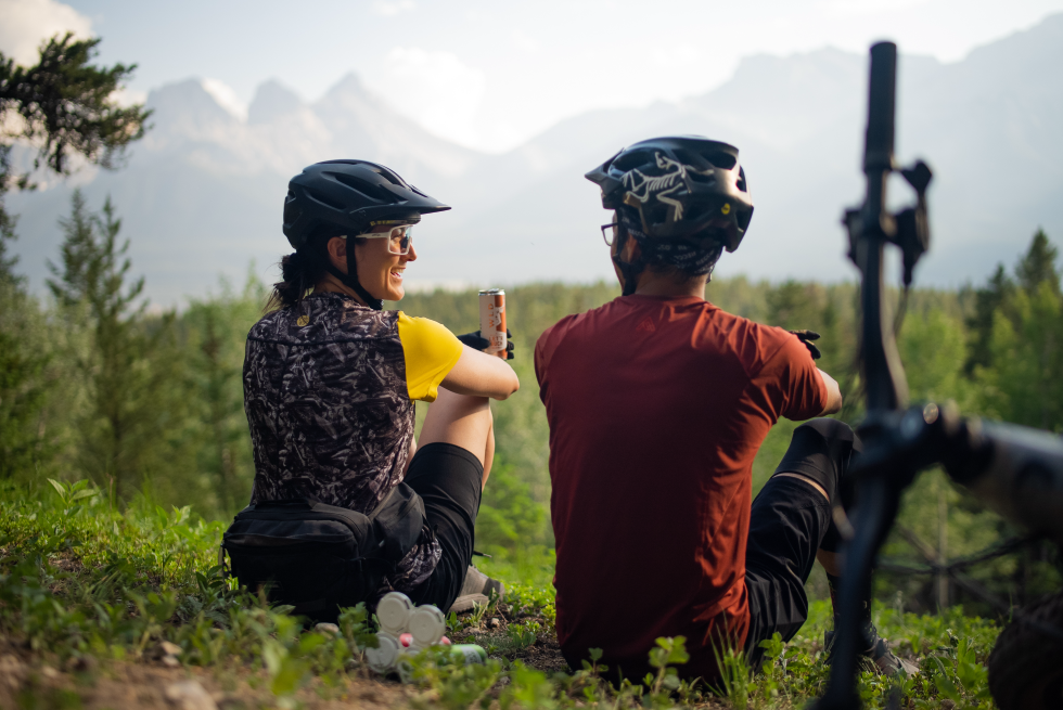 A woman and a man in mountain bike clothing sit infornt of an out of focus view of the Three Sisters and Ships Prow peaks above Canmore, AB. Seen from the back, she turns her head and smiles at him holding a canned cocktail form Wild Life Distillery. A bike lays on the ground next to them and between them is a 4 pack of canned cocktails.