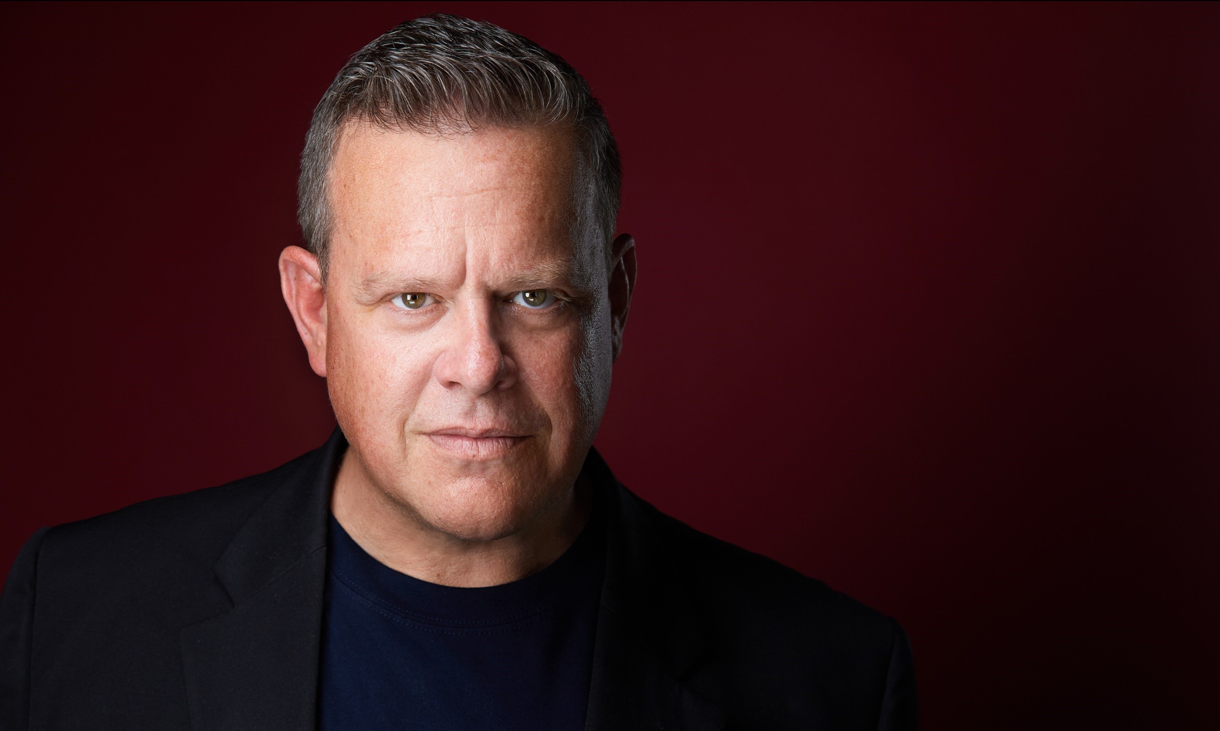 Man in dark clothing with short cropped blonde hair in front of burgundy backdrop 