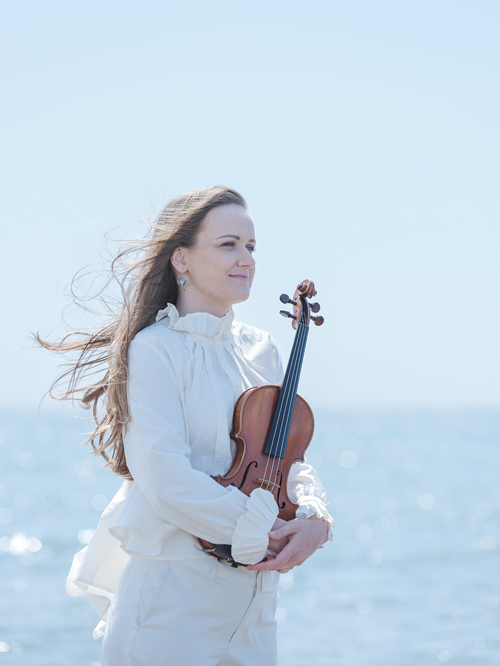 Woman standing in front of water in flowy clothes holding violin