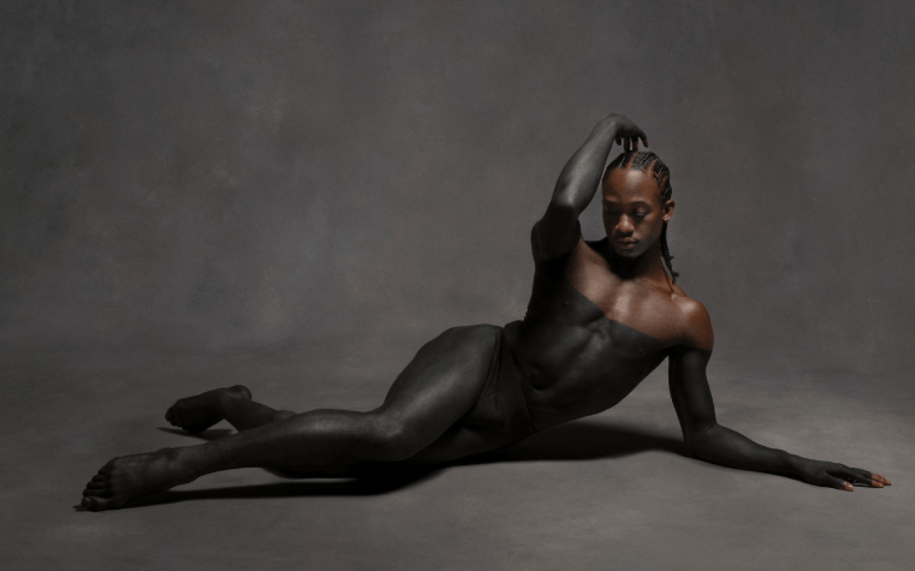 A dancer (Mykiah Goree) lies on the floor with fingertips lifted over their braids, gaze lowered. Their body is painted black to the shoulders, revealing brown skin above the paint against a gray background.