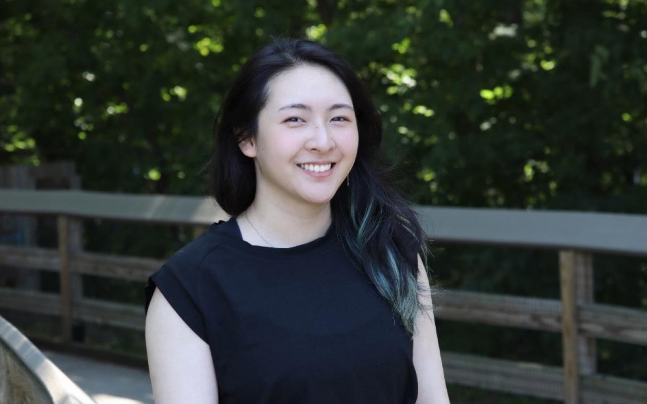 Smiling woman with dark hair posing by a wooden fence.