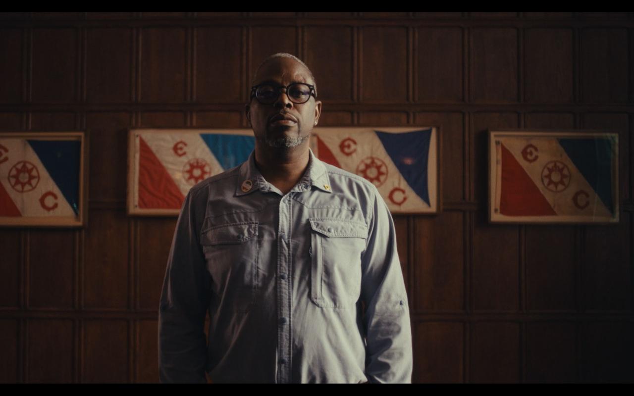 Dark-skinned man with glasses wearing a light blue shirt is standing in front of a blue white and red flag  hanging off a wood wall.