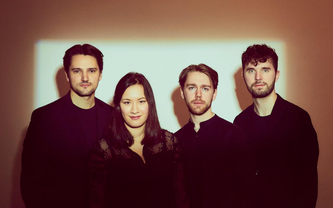 Four musicians dressed black, standing in front of a backlit wall