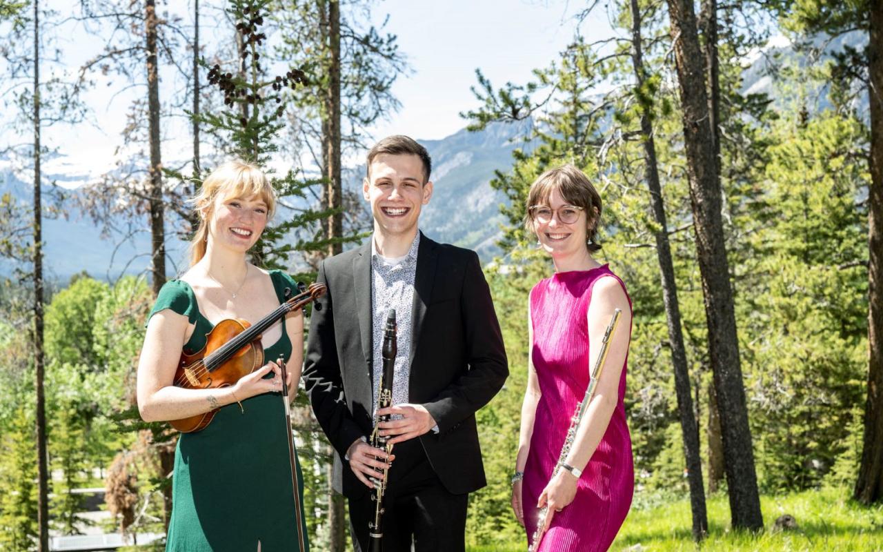 Woman in green dress holding a violin standing beside man in suit with a clarinet in front of him next to a woman in a pink dress holding a flute casually at her side