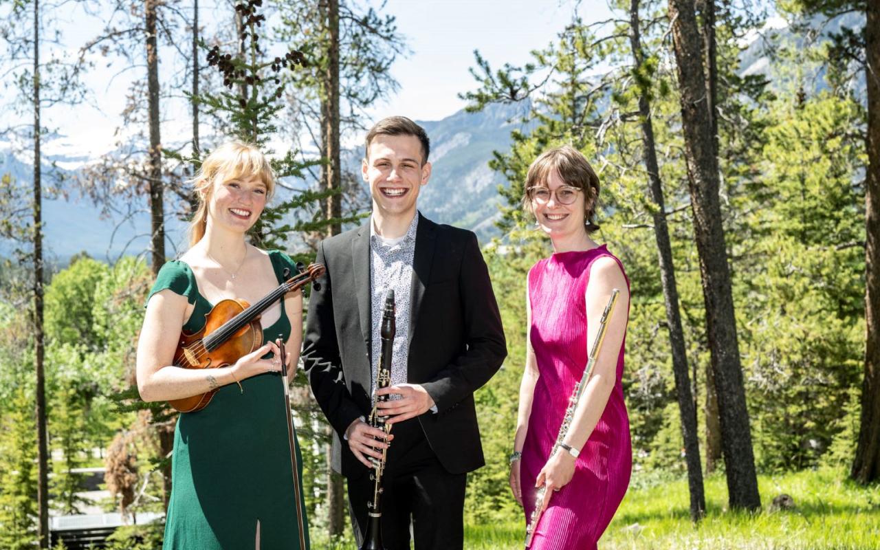 Woman in green dress holding a violin standing beside man in suit with a clarinet in front of him next to a woman in a pink dress holding a flute casually at her side