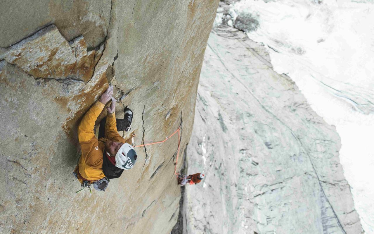 Man wearing yellow jacket climbing a big wall. Background shows a second man and glacier at the bottom. 